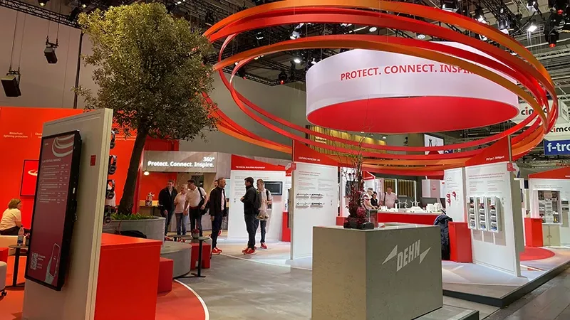 Exhibition booth with red and white design, several people, large red rings above the booth, and a tree in a red planter