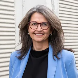 Portrait of a smiling woman wearing glasses, a blue blazer, and a black top, standing in front of a light-colored wall.