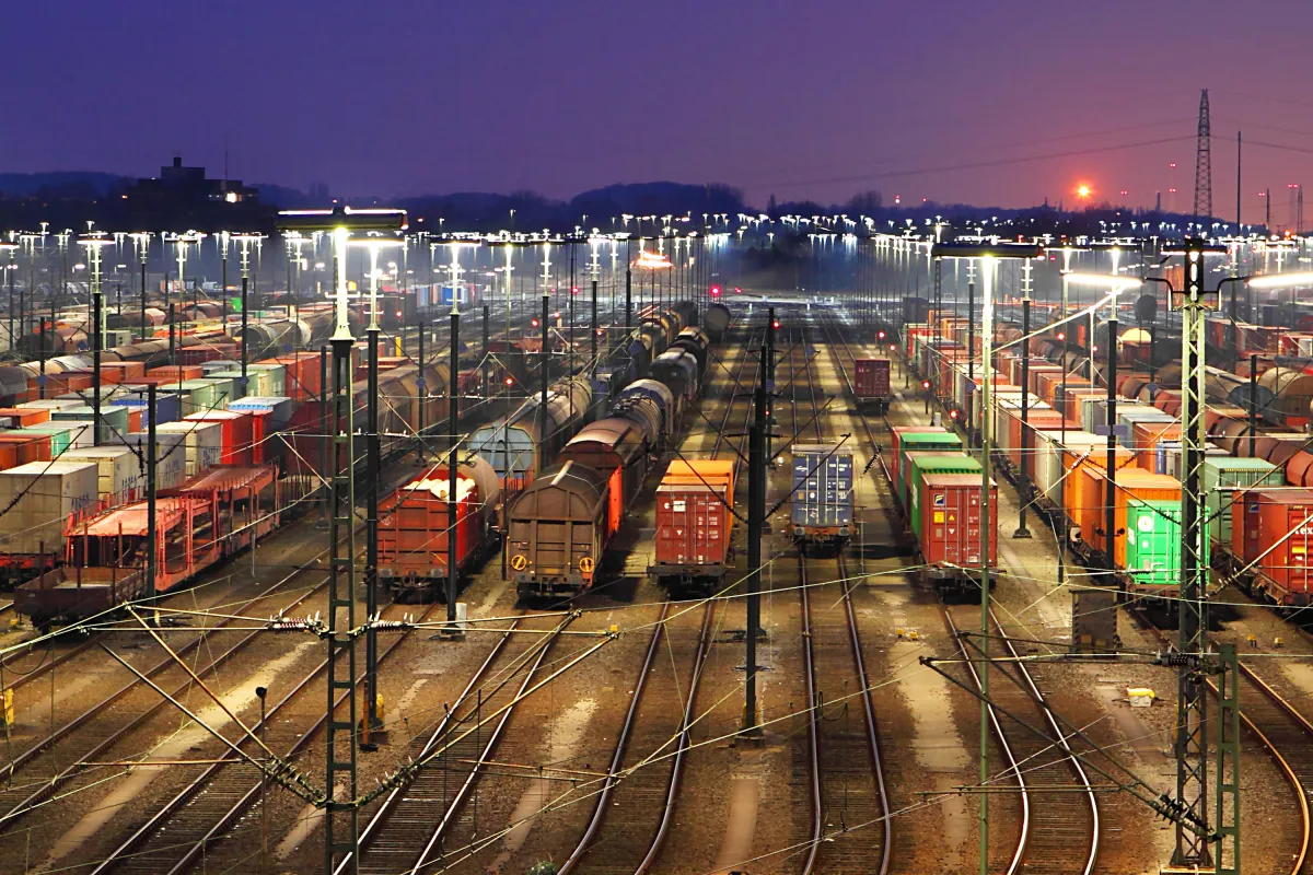 Numerous cargo trains and containers are lined up in a brightly lit train yard at dusk, with rail tracks converging into the distance and the sun setting on the horizon.