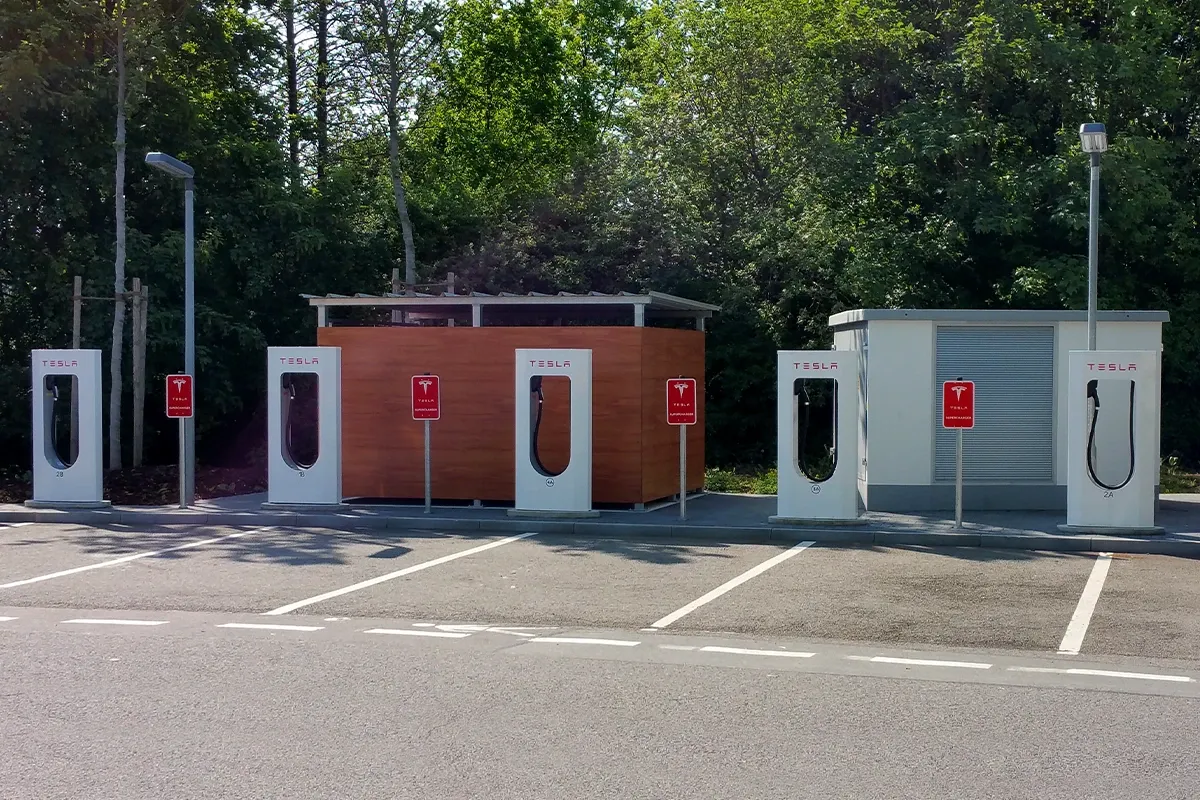 Tesla Supercharger station with multiple charging stalls in a parking area, surrounded by trees and small utility buildings.