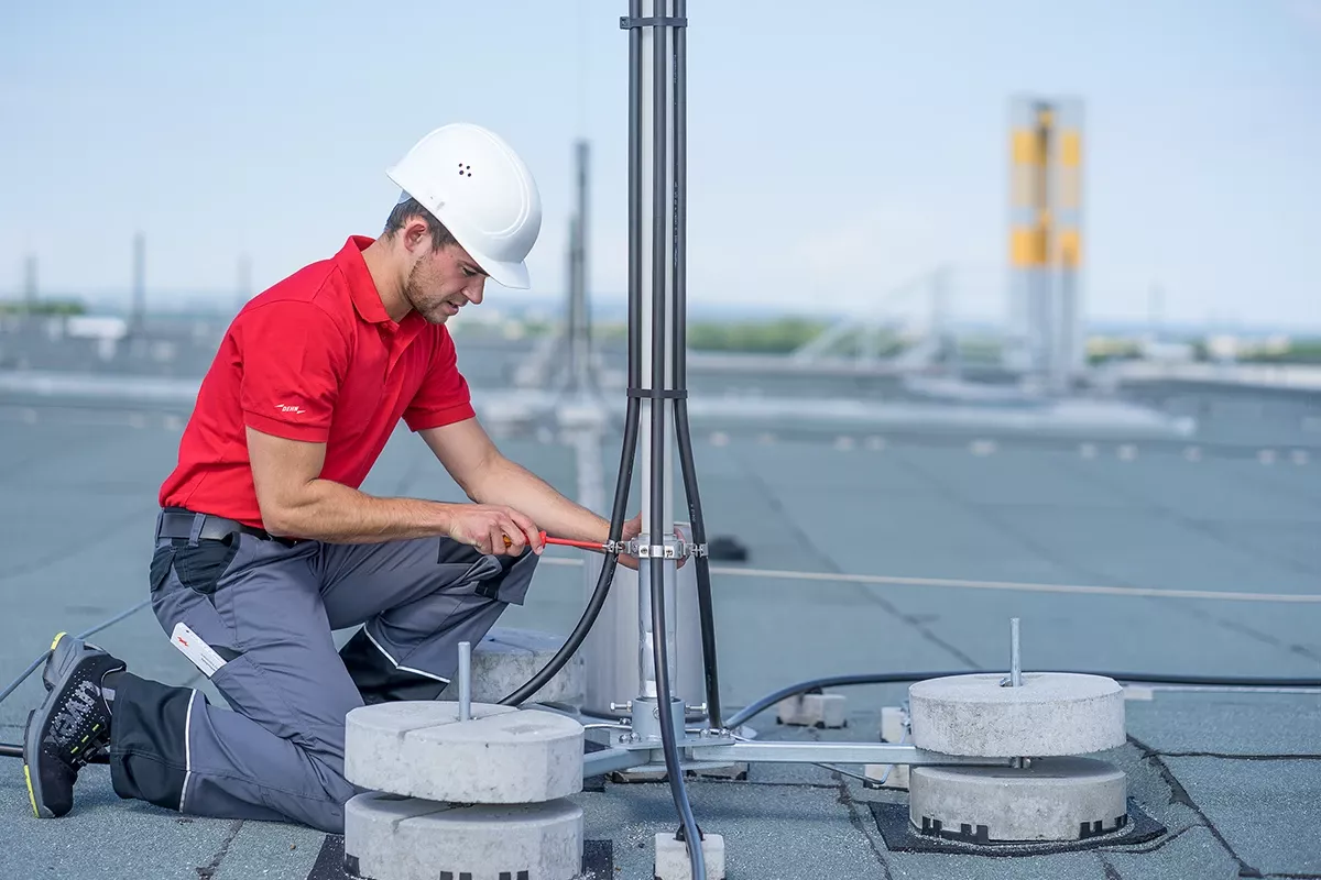 Technician in red shirt and white hard hat installing grounding cables on a rooftop lightning protection mast.